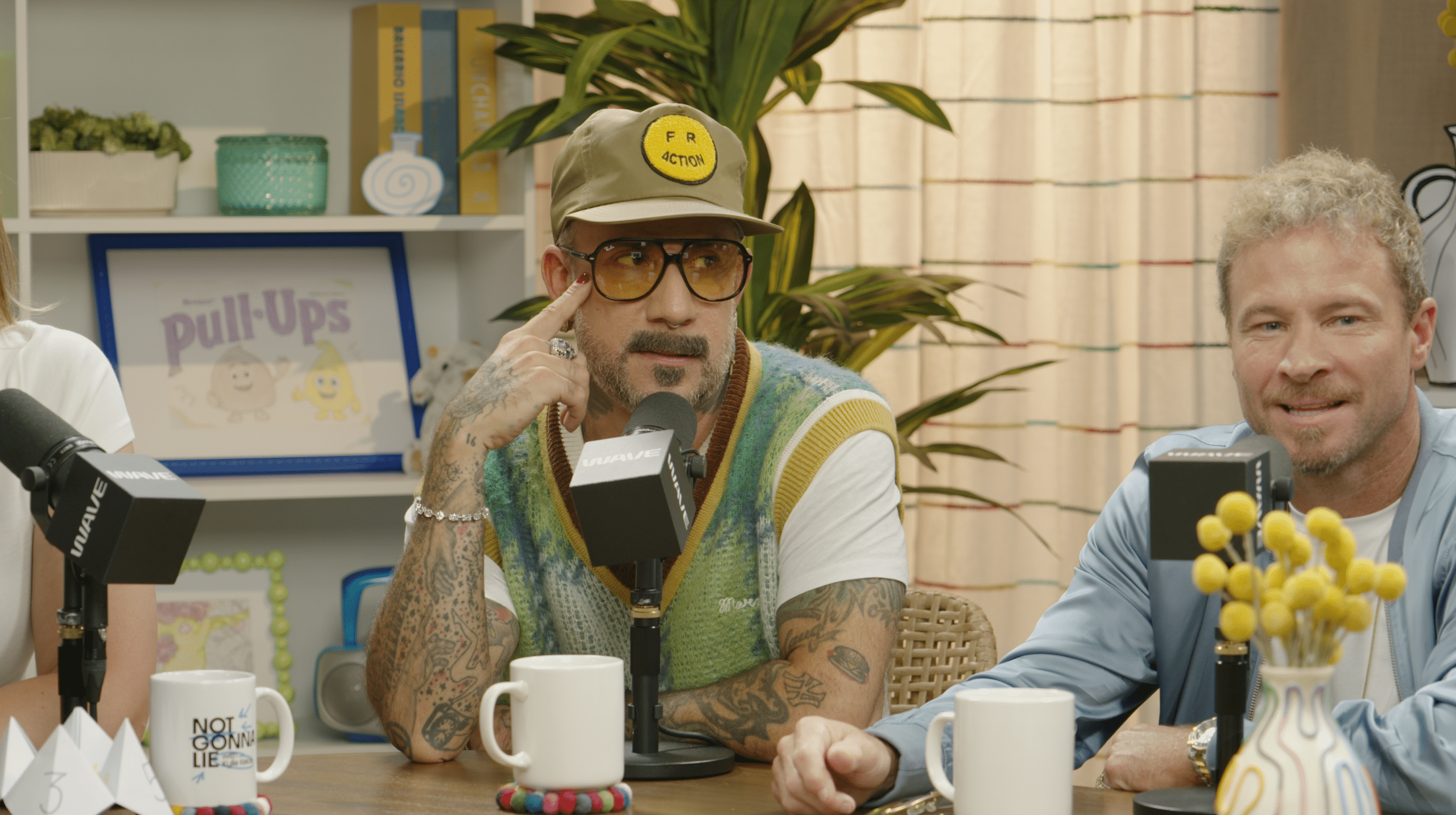 Two men sit at a table with microphones, one wearing sunglasses, a cap, and a colorful vest. The background has toys, books, and decor, suggesting a casual podcast or interview setting produced by ASL Studios.